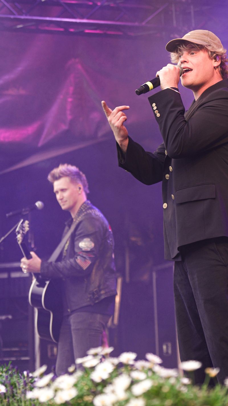 Nicki Jack med guitaren og Hjalmer ved mikrofonen på en scene med blomster foran